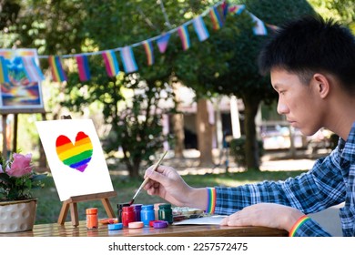 Asian teen gay wears rainbow wristband and holds paintbrush, drawing rainbow heart   in the park which decorated with rainbow flag and LGBTQ+ flagline, concept for special LGBT people events.