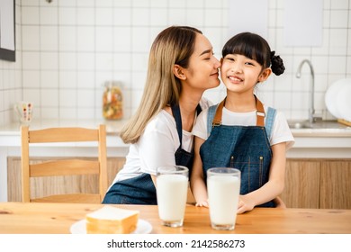 Asian Teen Girl Drinking Milk With Her Mom And Eating Bread, Sitting At Kitchen Table.Loving Mom Kissing Her Little Daughter In The Kitchen, Cheerful Family .