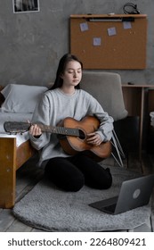 Asian teen girl playing guitar with laptop computer, learn to play with an online course in her room, sitting on the floor in casual clothes at home. Copy space - Shutterstock ID 2264809421
