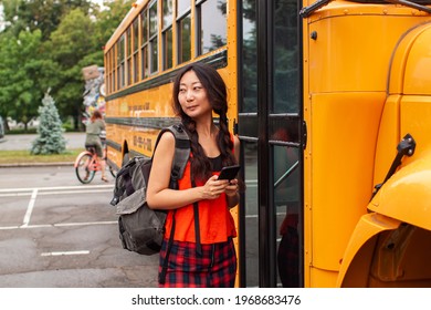 Asian Teen Girl Standing By A Big Yellow School Bus Door With Her Backpack.