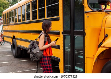 Asian Teen Girl Standing By A Big Yellow School Bus Door With Her Backpack.