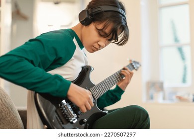 Asian Teen Guy Playing Electric Guitar Wearing Headphones Writing Song Sitting At Home. Boy Learning To Play Musical Instrument On Weekend. Teenager's Hobby And Music Talent. Selective Focus