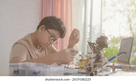 Asian teenage boy connecting wires to circuit board of robotic car with robotic arm at home, STEM education, robotics, and coding for young engineers.