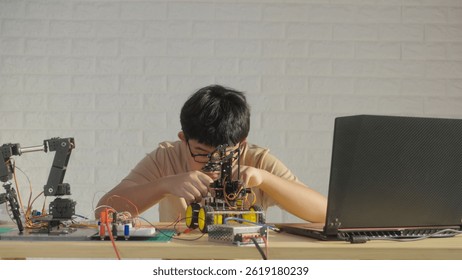 Asian teenage boy connecting wires to circuit board of robotic car with robotic arm at home, STEM education, robotics, and coding for young engineers.