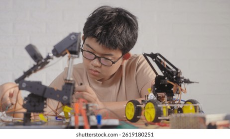 Asian teenage boy connecting wires to circuit board of robotic car with robotic arm at home, STEM education, robotics, and coding for young engineers.