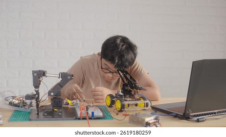 Asian teenage boy connecting wires to circuit board of robotic arm at home, STEM education, robotics, and coding for young engineers.