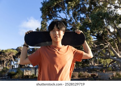 Asian teenage boy holding skateboard behind head, smiling outdoors in park. Youth, skateboarding, lifestyle, fun, leisure, urban, unaltered - Powered by Shutterstock