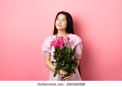 Asian Teenage Girl In Cute Dress Looking Romantic At Empty Space Logo, Holding Valentines Day Flowers Gift, Receive Boquet Of Roses From Lover, Standing On Pink Background