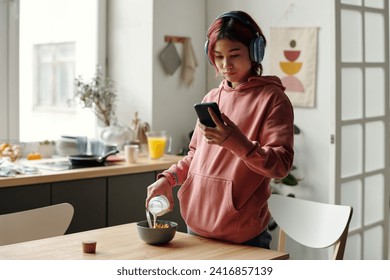 Asian teenage girl in headphones and pink hoodie pouring milk into bowl with cornflakes and watching online video on screen of smartphone - Powered by Shutterstock