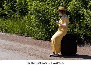 Asian teenage girl in a yellow dress, glasses and a hat sitting on a suitcase and looking for a navigator on her phone. The girl got lost. Ecotourism. Summer time. Holidays. Travel concept - Powered by Shutterstock