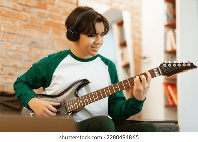 Asian Teenager Boy Learning To Play Electric Guitar Online Sitting At Laptop Computer At Home, Wearing Wireless Headphones. Modern Teen's Hobby Concept. Selective Focus On Guy - Powered by Shutterstock