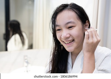 Asian Teenager Girl Cleaning The External Ear Canal With A Cotton Swab,smiling Young Woman In White Bathrobe,using Cotton Bud To Wipe Ears To Dry After Shower In Bathroom,removing Ear Wax,health Care