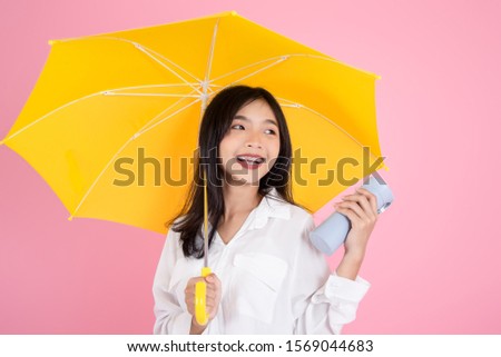 Asian teenager girl holding yellow umbrella during cold rainy day,drinks hot coffee to warm herself, pink background.  Autumn concept