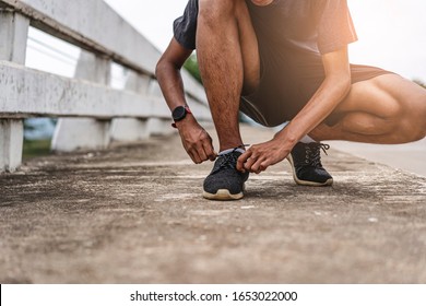 Asian Teenager Tying Shoe Lace After Running Along The Bridge, Exercising Or Practicing For A Marathon Race, Wearing Shirt, Short And Running Shoes, With Tree, Nature And Cloudy Sky In The Background