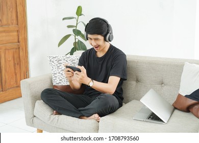 Asian Teenagers Play Games On Cell Phones While Relaxing On The Living Room Couch. Asian Teens Are Excited When Playing Games From A Cell Phone