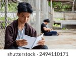 Asian teenboy in a dark jacket sits with his knees pressed against a metal fence panel and reading a daily notebook in a juvenile detention facility, awaiting further release, freedom and detention o