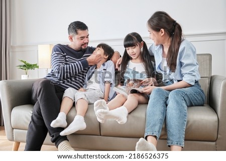 Asian Thai family, adult dad, mum, and children happiness home living relaxing activities and reading book together, leisure on sofa in white room house, lovely weekend, wellbeing domestic lifestyle.