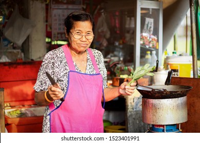 Asian Thai Local Street Food Vendor
Woman Avoid Sunlight In Shade And Cooking For Sale. Old Female Wear Glasses Aprons With Sunset And Blurred Small Carte Restaurant In Local Housing Estate Background