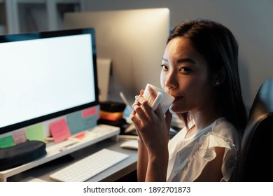 Asian Thai Woman Drinking And Sipping Hot Coffee From A Cup, Sitting Infront Of Computer At Office. Night Shift Time.