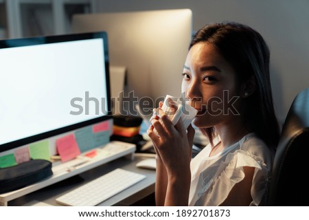 Asian thai woman drinking and sipping hot coffee from a cup, sitting infront of computer at office. Night shift time.