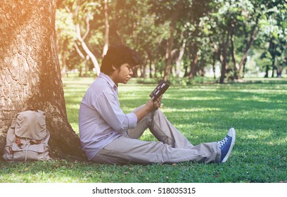 asian tired man resting and reading a book under a tree in a green park 