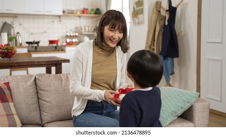 Asian Toddler Boy Walking Up To His Dad And Showing A Surprise Gift At Home. Happy Mother Embracing His Son For The Sweet Act