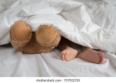 asian toddler feet beside teddy bear feet in white bed, sheet and pillow - toddler sleeping with teddy bear - Powered by Shutterstock