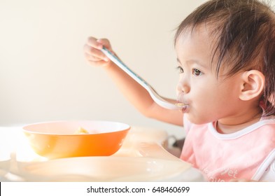 Asian Toddler Girl  Try To Eat Breakfast, Cereals Meal With Milk By Herself On Baby Chair In Morning