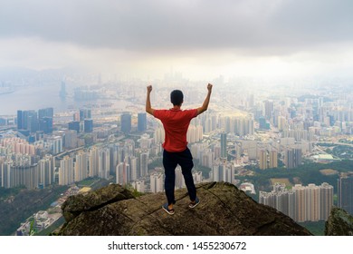 An Asian Tourist Man Hiking And Looking At City On Rock, Mountain Hill In Hong Kong Downtown In Adventure Concept During Travel Trip, Vacation, Or Holidays. Skyscraper And High-rise Buildings View.