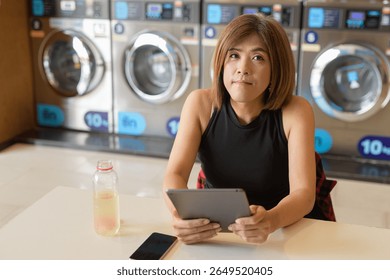 Asian transgender non-binary person sitting in laundromat laundry shop using tablet computer