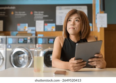 Asian transgender non-binary person sitting in laundromat laundry shop using tablet computer