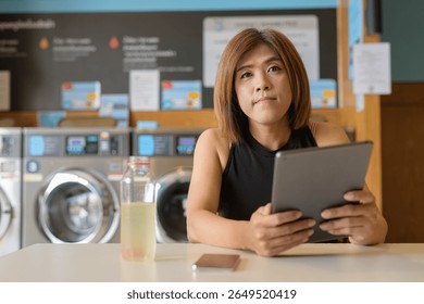 Asian transgender non-binary person sitting in laundromat laundry shop using tablet computer