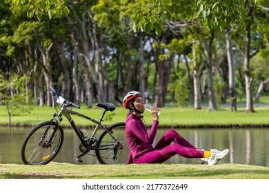 Asian transgender woman in sportswear taking a rest and drinking water for rehydration after cycling with sport bicycle around public park by the lake