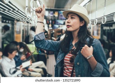 Asian Travel Woman Taking Underground Metro Standing Holding Handle On Train. Female Backpacker In Subway By People Sitting Reading Resting On Seat. Girl Waiting For Her Destination Arrived In Japan.