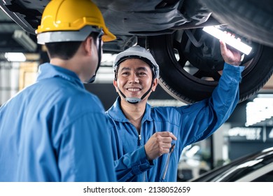 Asian Two Automotive Mechanic Men Wear Helmet Work In Mechanics Garage. Team Of Vehicle Service Manager Male Worker Look Under Car Condition, Check And Maintenance To Repair Engine Machine In Workshop