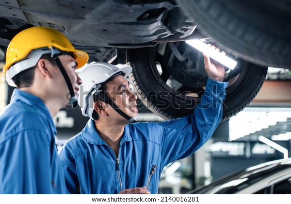 Asian two automotive mechanic men wear helmet\
work in mechanics garage. Team of vehicle service manager male\
worker look under car condition, check and maintenance to repair\
engine machine in\
workshop