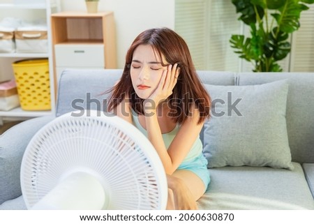 asian unhappy woman is sitting in front of working fan suffering from summer heat at home