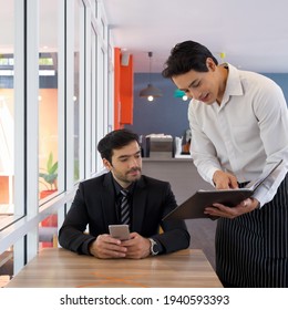 Asian Waiter In Apron Pointing Finger At Restaurant Menu. Recommend Chef Choice Menu To The Customer. Young Businessman In Suit Listening To The Waiter While Holding Mobile Phone.