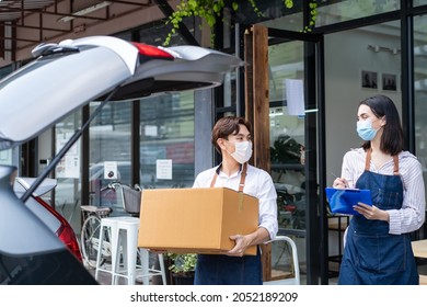 Asian Waiter Loads Parcels Into Car With Waitress Check On Clipboard. Cafe Business Owner Couple Wear Mask, Work In Coffee Shop During Covid-19 Pandemic . Delivery Purchased Transportation Concept.