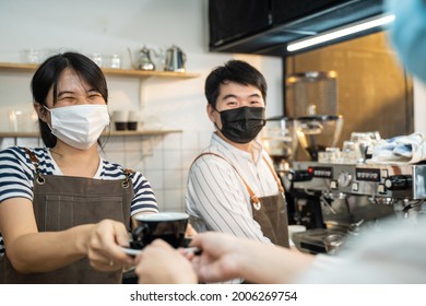 Asian Waitress Hand A Cup Of Coffee To Customer With Two Hand At Cafe. Business Owner Barista Woman In Apron Wear Mask Due To Covid19 Pandemic, Work In Restaurant Serve Occupation Service Young Girl.