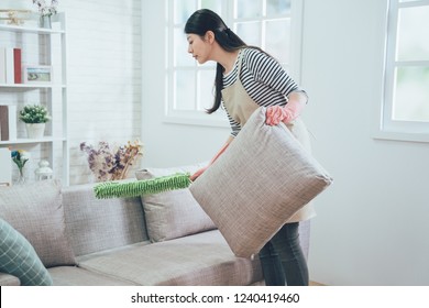 Asian Wife Using Green Feather Duster Carefully Cleaning The Sofa. Young Female Housekeeper Taking Up Pillow Dusting The Couch In The Cozy Living Room At Apartment. Lady Doing House Chores In Apron.