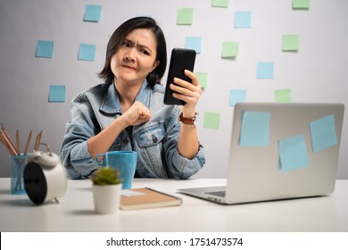 Asian Woman Angry Looking At Smart Phone Reading News At Home Office. WFH. Work From Home. Prevention Coronavirus COVID-19 Concept.