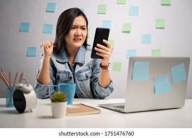 Asian Woman Angry Looking At Smart Phone Reading News At Home Office. WFH. Work From Home. Prevention Coronavirus COVID-19 Concept.