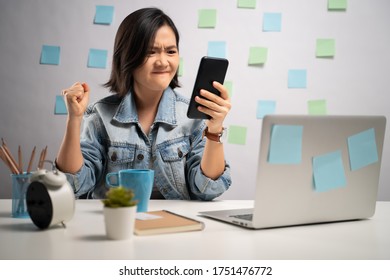 Asian Woman Angry Looking At Smart Phone Reading News At Home Office. WFH. Work From Home. Prevention Coronavirus COVID-19 Concept.