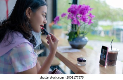 Asian Woman Apply Red Lipstick On Lips At Cafe Using Smartphone As Mirror, Girl Have A Drink And Make Up At Wooden Table In Coffee Shop