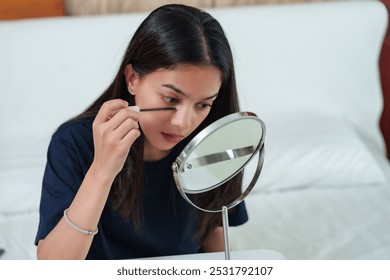 Asian woman applying mascara while sitting on bed, focused on enhancing eyelashes using a mirror for precision. Quiet bedroom environment with beauty tools laid out, part of morning makeup routine.