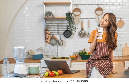 Asian woman in apron sitting on kitchen counter using smartphone smiling while browsing recipes or watching videos, modern home kitchen, fresh fruits and vegetables, cooking concept, healthy lifestyle - Powered by Shutterstock