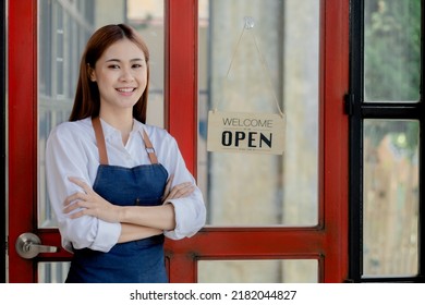 Asian Woman In An Apron Stands Holding A Restaurant's Menu In Front Of The Door With A Sign Opening A Shop, An Opening Of A Small Restaurant, A Woman Running A Business. Operating A Small Restaurant.