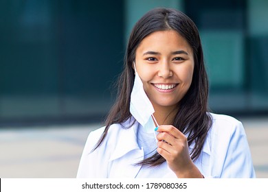 Asian woman, attractive ethnic girl takes off the medical mask from her face, happy with a smile looks at the camera. in a white shirt outside the business center.
