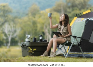 An Asian woman backpacker sitting and holding a digital tablet while camping in a park. - Powered by Shutterstock